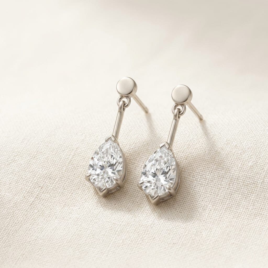 Gota earrings: two pear-cut diamonds in white gold claws, side by side on ivory linen